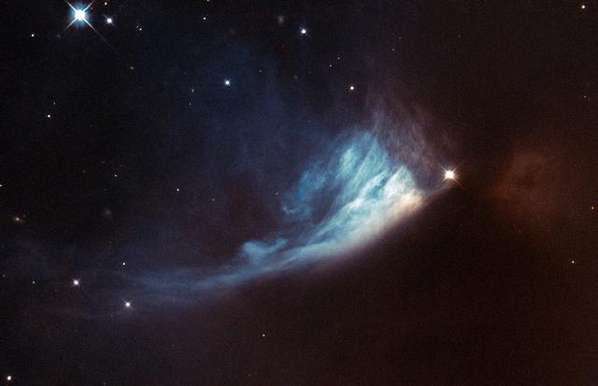 Astrophotography image from the Hubble Space Telescope of  PV Cephei and Gyulbudaghian’s Nebula. A bright star is in the center of the right side and a fan of blue, green, and white gas seems to be flowing behind the bright star across the image. Other stars and bright objects are scattered throughout the dark sky.