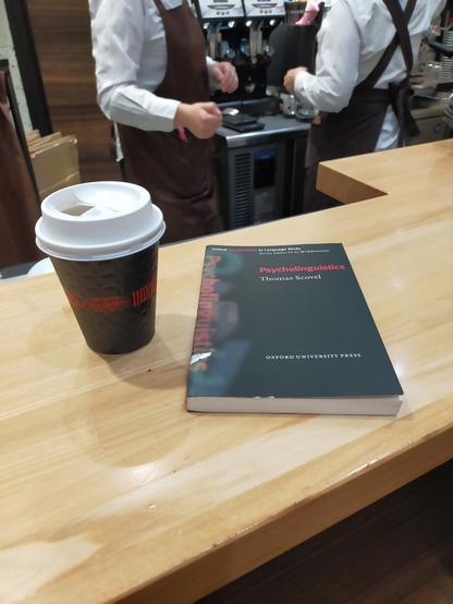 The small black paperback book with the title PSYCHOLINGUISTICS in red is on a light brown wooden counter. To tye right is a black paper coffee cup with a red band in the middle with the unreadable name of the coffee shop. It has a white plastic lid. Behind the counter we can see 2 headless baristas in brown aprons & white dress shirts working in front of an espresso machine