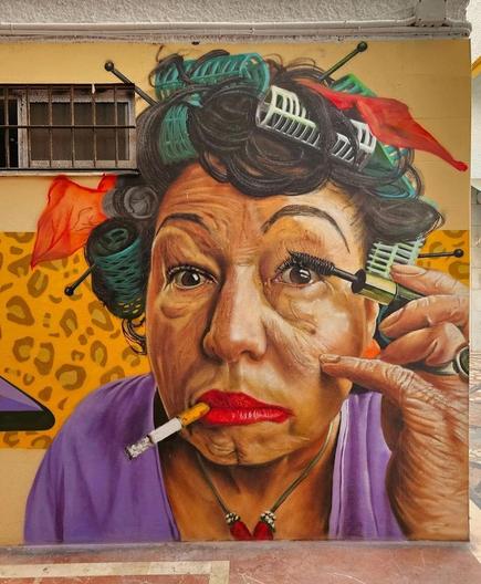 Streetartwall. An unusual portrait of a lady putting on make-up was sprayed/painted on a long street wall. The elderly lady is wearing a purple T-shirt, a necklace and green curlers in her black/gray hair. She is depicted larger than life and appears to be looking into a mirror, although this is invisible to us. In any case, we see her with a cigarette in the corner of her deep red lips while she applies mascara to her eyelashes with a mascara brush. And the feeling of disturbing something creeps up on us.