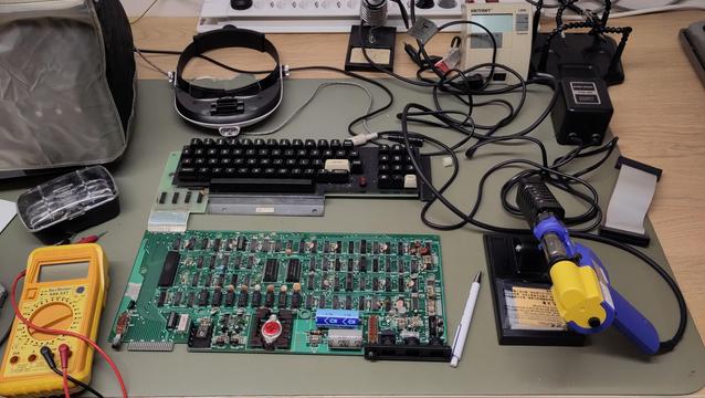 A green circuit board with a keyboard attached through a ribbon cable on a desk, flanked by a yellow multimeter on the left, magnification goggles on top and a blue/yellow desoldering gun to the right, along with a tangle of wires and a soldering iron. There's also a TRS-80 under a beige cover.