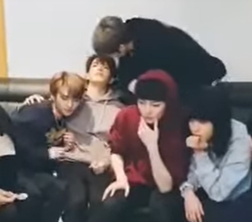 several skz members on a couch. Hyunjin snuggles up against Seungmin's side, while Lee Know leans over from the back of the couch to get in Seungmin's face