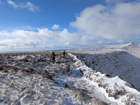 Two people walking along a snowy ridge with flat snow covered heath on one side and a precipitous drop on the other.
