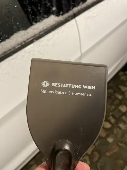 Das Bild zeigt eine dunkle, rechteckige Eisschaufel, die an einem weißen Auto gehalten wird. Die Schaufel ist mit dem Logo und dem Namen „Bestattung Wien“ und dem Slogan „Mit uns kratzen Sie besser ab.“ bedruckt.  Das Auto ist teilweise mit Schnee oder Eis bedeckt. Der Hintergrund ist unscharf, zeigt aber einen Teil eines gepflasterten Weges.