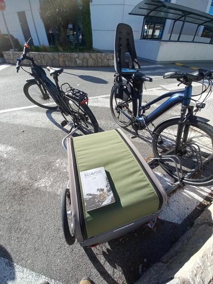 Un exemplaire du livre "Bikepunk" de Ploum, posé sur la cariole attachée à mon vélo. On distingue un autre vélo avec un siège bébé sur la droite.
