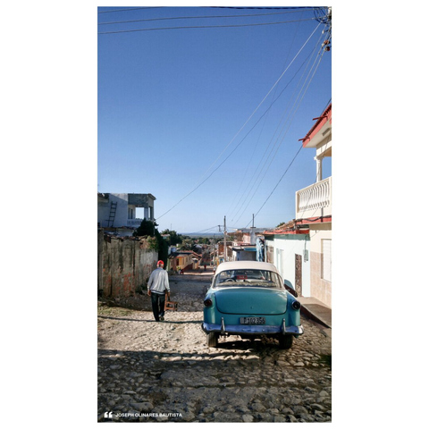 A classic turquoise car sits parked on a cobbled street in Trinidad, Cuba. An elderly man walks slowly ahead, carrying a wooden chair—perhaps to sit and chat with an old friend just down the road—against a backdrop of pastel houses and a view of the sea in the horizon. The serene morning scene captures the town’s unhurried rhythm beneath a crisp, cloudless sky. Photo taken by Joseph Bautista.