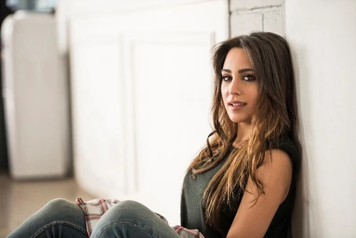 A promotional image of Mayssa Karaa, a slender brunette woman with darkly-accented eyes resting against a white wall in a well-lit room.