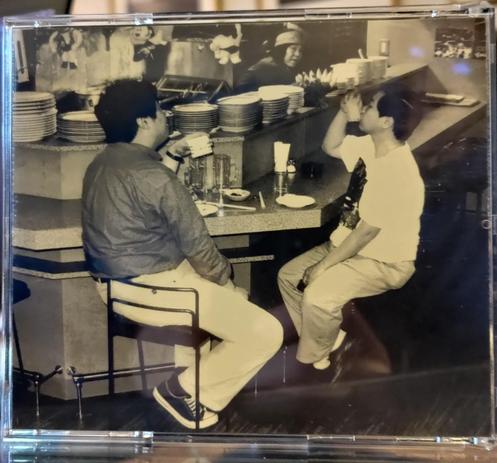 jewlcase CD cover. backside. black and white photo:Toshiji Mikawa and Fumio Kosakai sitting and drinking, seen from the side. woman behind the counter, with stacks of plates before her.