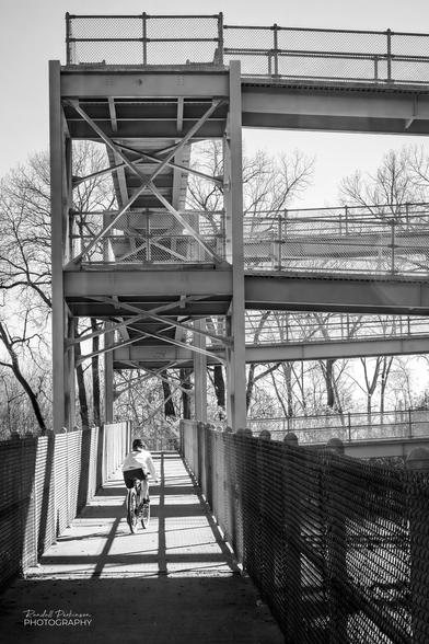A cyclist heads up a square shaped, metal framed bike and pedestrian ramp providing access to cross a large river bridge out of the picture.