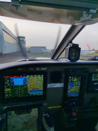 Instrumente panel of a light aircraft with multiple computer screens, displaying primary flight instruments, a map, and engine parameters; a tablet with flight navigation software is mounted on the yoke. Hangars and the spinning propeller can be seen through the windshield.