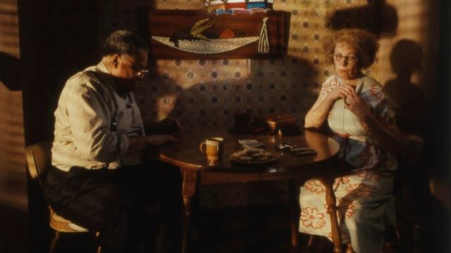 The image depicts a dimly lit room featuring two individuals seated at a small wooden table. The person on the left is a man wearing a white shirt and glasses, focusing on something in front of him. The person on the right is a woman in a light blue dress with orange floral patterns, also wearing glasses, with her hands clasped together. The table between them holds a yellow mug, a plate with cookies, and some folded papers or small cards. The background is adorned with retro-patterned wallpaper and a wooden wall hanging designed like a stylized sailboat.