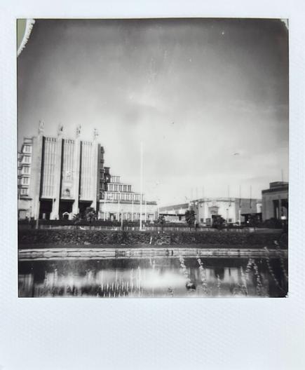 Black and white, Polaroid photo: a building built for the 1935 international exhibition towers over, and is reflected in, an artificial lake at the fairground in Brussels.