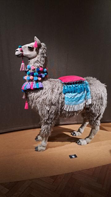 Art work, a model of a grey fluffy llama with a multicoloured cloth saddle and headless.