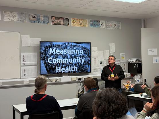 A presentation taking place at BarCamp London. The speaker, Alan Pope, is standing in a classroom-like setting, wearing a black shirt and a red lanyard. Behind him, a large screen displays the title "Measuring Community Health" in bold letters, overlaid on a black-and-white image of a crowd. Several attendees are seated, listening attentively, with some using laptops. The room features posters related to mathematics on the walls and some office equipment in the background.