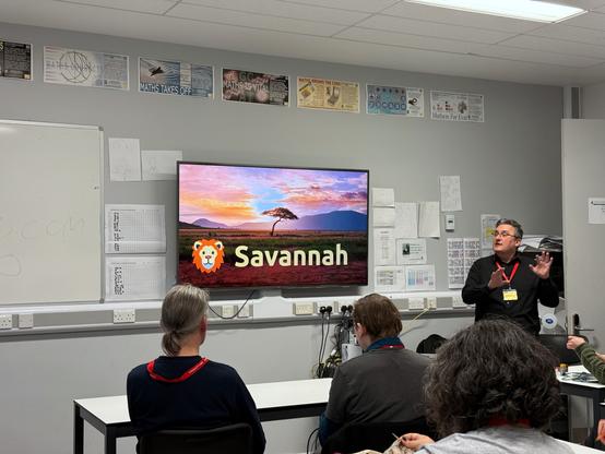 A presentation at BarCamp London by Alan Pope. The speaker, wearing a black shirt and a red lanyard, gestures with his hands while addressing the audience in a classroom-like setting. On the screen behind him is a vibrant image of a savanna at sunset, with a tree and mountains in the background. The title "Savannah" is displayed alongside a lion cartoon logo. Several attendees are seated, watching the presentation, with some taking notes or using laptops. The room features posters related to mathematics on the walls and office materials in the background.