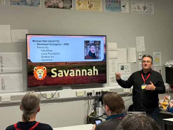 Alan Pope presenting at BarCamp London in a classroom-like setting. On the screen behind him is a slide featuring the title "Savannah" with a lion cartoon logo, alongside a profile for Michael Hall, labeled as "Developer Evangelist - ARM." The slide lists Michael Hall’s previous roles at InfluxData, Linux Foundation, Endless Inc., and Canonical, with a photo of him included. Alan Pope, wearing a black shirt and a red lanyard, gestures as he speaks to the seated audience, some of whom are taking notes or using laptops. Posters about mathematics decorate the walls.