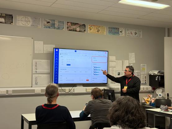 Alan Pope giving a presentation at BarCamp London in a classroom-like environment. He is pointing at a large screen displaying a software interface with the title "Phil Clifford" and a section labeled "Opportunities." The screen appears to show a CRM or task management tool with various details and metrics. Alan Pope, wearing a black shirt and a red lanyard, engages with the seated audience, who are attentively watching or taking notes. The room features posters related to mathematics on the walls and some office equipment in the background.