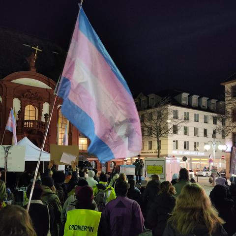 Versanmlung von Menschen auf dem nachtdunklen Markplatz in Mannheim. Vorne auf einer Plattform steht ganz klein eine Person mit einem Mikro. Prominent im Bild eine wehende Trans Pride Flag.