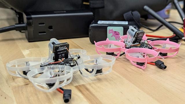 Two tiny racing drones resting on a table top, one with a clear frame, clear props, and a clear camera mount. The other with a pink frame, clear props, and a clear camera mount.