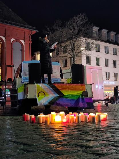 Kundgebung auf dem nachtdunklen Marktplatz in Mannheim. Eine Person auf einer Redner*innentribüne. Die Tribüne ist mit einer Progressive-2-Pride-Flag geschmückt, im Hintergrund eine Trans Pride Flag.Auf dem Boden vor der Tribübne brennen Kerzen.
