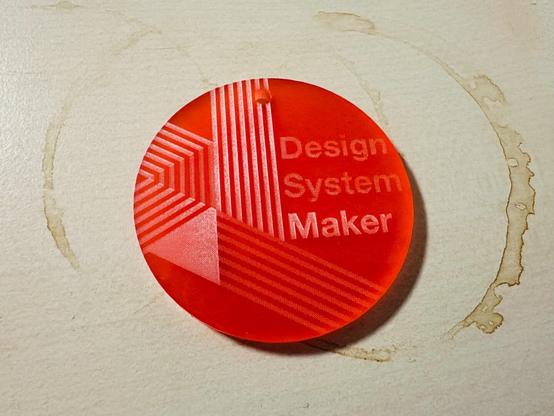 Un objet rouge en plastique ou plexi avec des gravures dessus. Il est écrit « Design System Maker ». Les gravures et écritures semblent avoir été faites au laser. En haut il y a un trou pour passer par exemple un anneau de porte-clés. L’objet est posé sur une surface faisant penser à un bureau avec de nombreuses traces de tasse de café dessus.