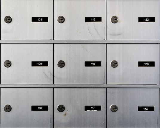 The image depicts the interior of a mailroom filled with metal mailboxes of various sizes. The mailboxes are lined up in neat rows along the walls, creating a grid pattern under bright, artificial light. The room has a monochrome appearance, emphasizing the contrast and shadows.