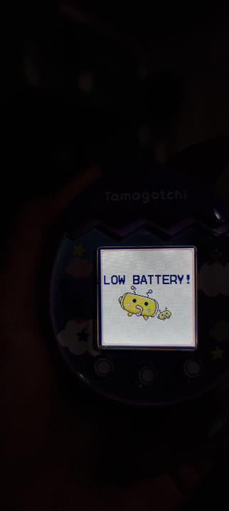 A photo of a person holding a tamagotchi pix in the dark. On the screen it says Low battery! at the top and has a picture of one big and one small battery shaped yellow tama with antenna.