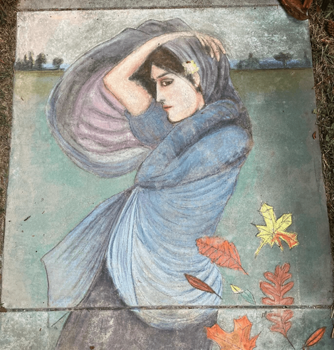 A chalk drawing on pavement, of a woman with pale skin and dark hair and eyes, wrapped in a large blue shawl, one hand behind her neck, one hand over her head; there are some autumn leaves in the foreground).