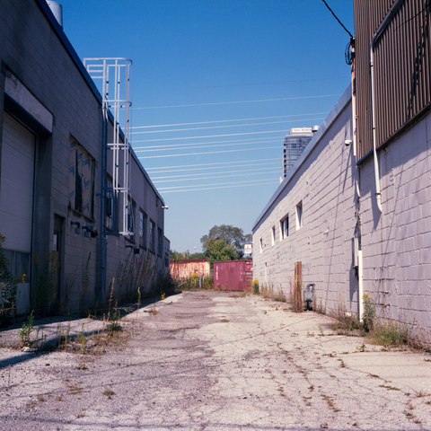 The patchwork pavement driveway between two low industrial buildings with two shipping containers at the end, the buildings are vacant and showing neglect and one of the containers is showing its decay. A score of transmission lines cut diagonally across the sky between them, gleaming in the sun.