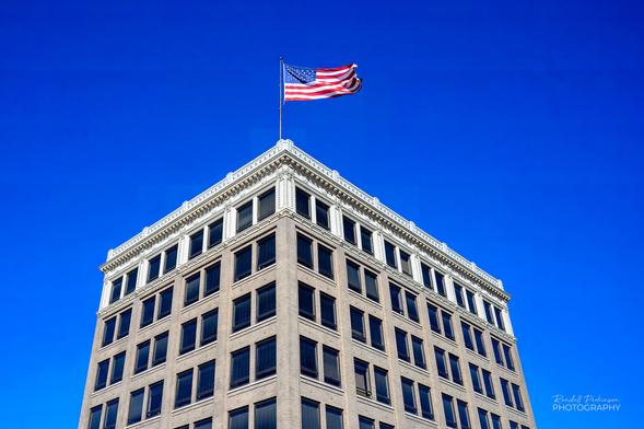 An older multi-storied office building with a large USA flag flying from a pole on the roof set against a clear blue sky.