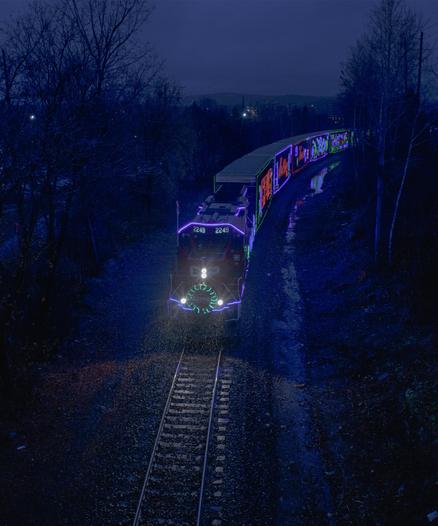 On a dark and rainy night, an illuminated Christmas-themed railroad train travels forward towards the viewer.  The lights on the side of the train's boxcars are reflected in puddles near the train tracks.