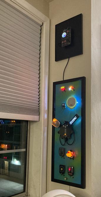 Photo of two frames hanging on a wall next to a window. The upper frame is smaller and black, showing the Supercon badge from 2023. The lower frame is larger and painted blue and green and has eight illuminated SAOs arranged around the Supercon badge from 2024 in the center. Detailed annotations are noted on my blog post.