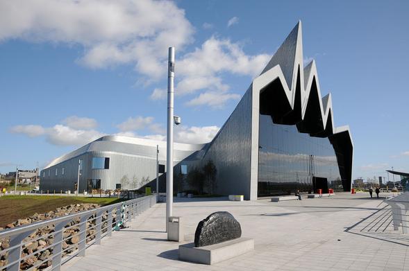 The Riverside Museum on the River Clyde in Glasgow. The view shows the exterior of the museum with a paved area in the foreground and the curved metal-clad body of the museum on the left of the structure. On the right is the spiky frontage overhanging its glass face which seems black and is highly reflective. The sky is mainly blue.