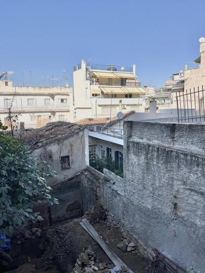 In the background, against blue sky, sunlit apartment blocks, 3-5 storeys, dating from the mid to late 20th Century. Long balconies, awnings, flat roofs.
In the foreground, seen from above the deeply shaded empty lot where a 19th to early 20th Century house has collapsed of neglect. A tree grows in it. A surviving, decrepit building of that period bounds the further side of the earthen space. To the right, two newer flat-roofed buildings do, one with a lushly planted covered veranda recessed into the wall and looking towards the tree.