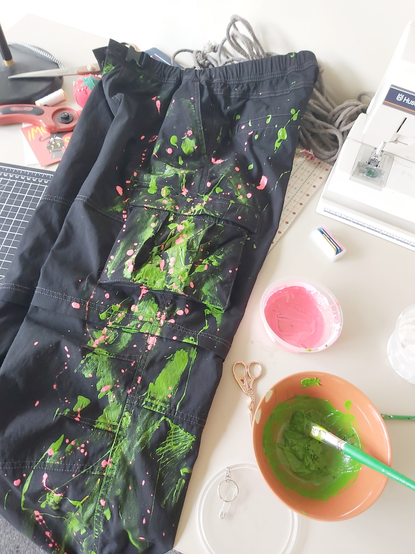 pair of black cargo pants with green and pink paint splatters, laid out on a desk with materials nearby: paintbrush, paint, thread scissors
