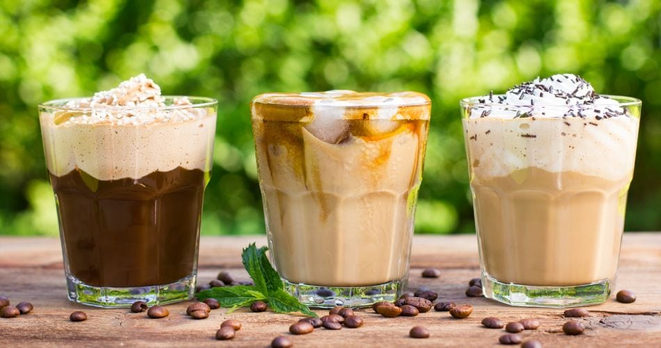 Foto van 3 luxe koffie's, elk met romige schuimlaag erop en elk met een net verschillende kleur, op een houten tafel met losse koffiebonen erop, met een out-of-focus groene achtergrond.