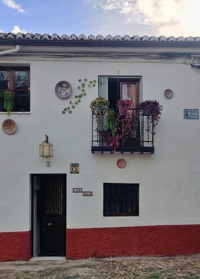 Fachada de una casa en el Albaycín. Hay un balcón con plantas y platos colgados alrededor. La puerta de la calle está entreabierta.