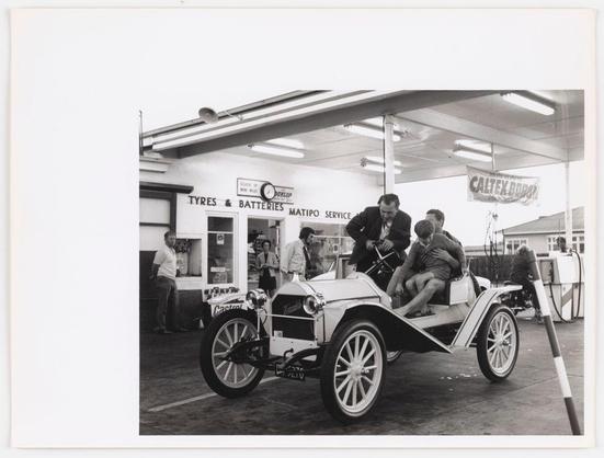 The image depicts a vintage automobile at what appears to be an early 20th-century fuel station. The car, with its open-top design and exposed engine components, is reminiscent of the Model T Ford era vehicles.

Inside the car are several individuals: one driver wearing glasses, another in casual attire holding onto a part of the vehicle's structure, while two passengers sit near the rear; both appear to be dressed for leisurely travel. There's also an individual seated outside on a chair next to the fuel pump attending to their own business.

Behind them is the station with various signs such as "TYRES & BATTERIES MATIPO SERVICE" and a banner reading "CALTEX BOILER," suggesting that this photo was taken during a time when Caltex, now known as Mobil, distributed oil products. The presence of these individuals suggests social interaction or perhaps travel from one location to another.

Overall, the scene captures a slice-of-life moment in urban life history, possibly Wellington's cityscape as noted by City and town life, Wellington. It reflects on historical modes of transportation and hints at everyday activities during that period within an early gas station environment, illustrating how fueling stations served not only vehicles but also community gatherings or personal breaks away from vehicle operation duties.

The photograph is in black-and-white which adds a timeless quality to the image, emphasizing the contrast be [...]