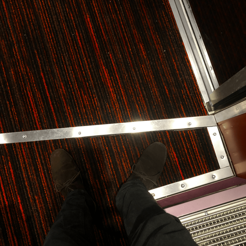 Looking down at a red and black thin striped carpeted floor divided by silver rectangular strips of metal. At the bottom on the frame two feet in dark brown suede shoes are visible. At the very bottom right one can see a metal grate and two coloured metal strips, one light pink, one dark pink.