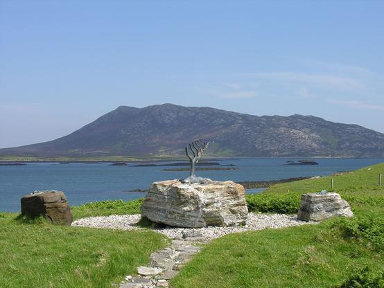 A view across Loch Euphort on North Uist in the Western Isles. The image shows the view on a sunny day, with a sculpture of what looks like a branch of a tree on a rock in the foreground. There is a stony area around the sculpture with a path leading to it. In the background is the loch, complete with islets. In the distance a rocky hill rises from the far shore.