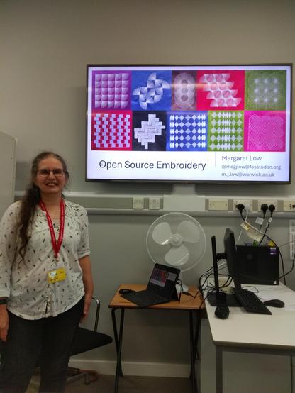 Margaret standing in front of a display screen, showing the title of the presentation "Open Source Embroidery" along with some pictures of stitched patterns.