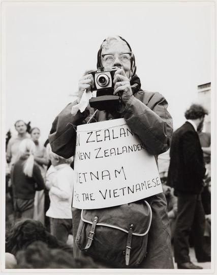 The image is a black and white photograph of an individual holding up a vintage camera to their eye, presumably taking a photo. The person wears glasses and has headphones or ear protection around their neck. They are dressed in layers with what appears to be a jacket over a hooded garment.

In front of the photographer's chest, there is a handwritten sign that reads "NEW ZEALANDER NEW ZEALANDER VIETNAM VIETNAMESE FOR THE VIETNAMESE." This suggests that they might have been participating in some form of protest or demonstration related to Vietnam. The background shows other individuals and what seems like an outdoor setting, indicating the presence of a group event.

The photograph is part of a series titled "Notes on the country I live in" by Ans Westra, as indicated at the source link provided. It captures a moment that likely relates to anti-war sentiment or solidarity with Vietnamese people during the Vietnam War era.