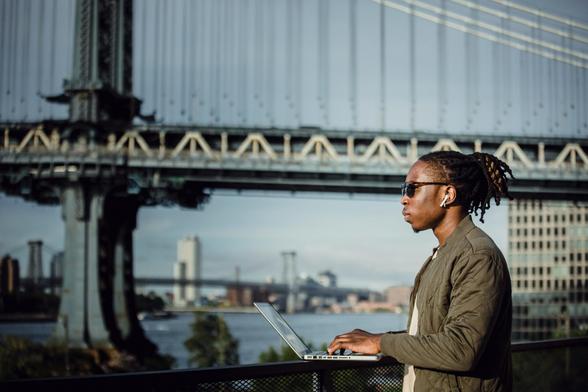 A photo of a person standing and typing on their laptop outside with bridges in the background (Photo by Ketut Subiyanto on Pexels.com)