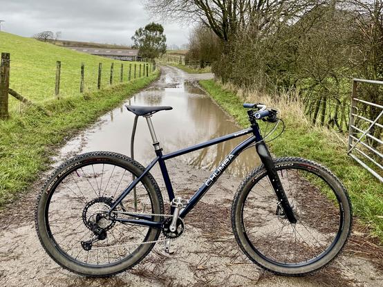 Dark blue mtb stood on a muddy lane in front of a puddle.
