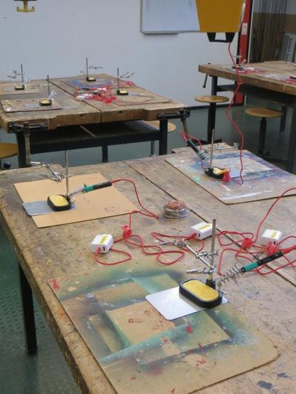 Mehrere Arbeitstische, welche für einen Lötworkshop vorbereitet worden sind. Zu sehen sind Lötunterlagen, Lötkolben (Pinecil v2), USB-Steckernetzteile , "dritte Hände" und Lötspitzenreiniger.