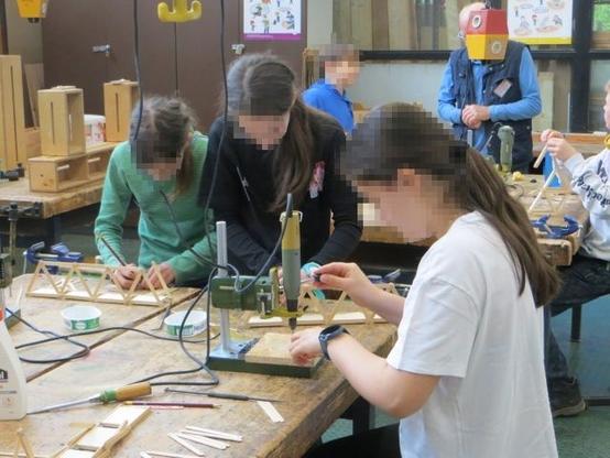 Kinder arbeiten mit Bohr- und Fräsgeräten in Bohständern an Werktischen. Sie bauen eine Fachwerkbrücke aus Holz.