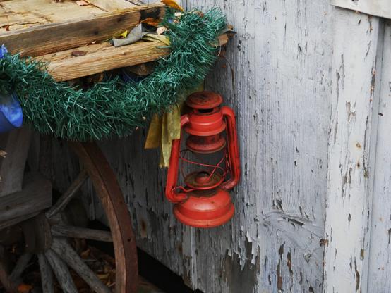 Ein Stilleben. Man sieht eine alte, rote und rostige Petroleumlampe ohne Glas und docht, welche an der Kante eines offensichtlich alten Wagens mit Holzrädern hängt, der an einer Holzwand steht. Die graue Farbe von der Holzwand bröckelt gerade ab.