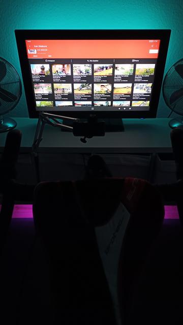 Vor einem Fahrrad im Smarttrainer steht ein Fernseher und 2 Ventilatoren auf einem niedrigen Regal. Die Wand hinter und der Boden unter dem Regal werden mit LED-Lichtern farbig erhellt.