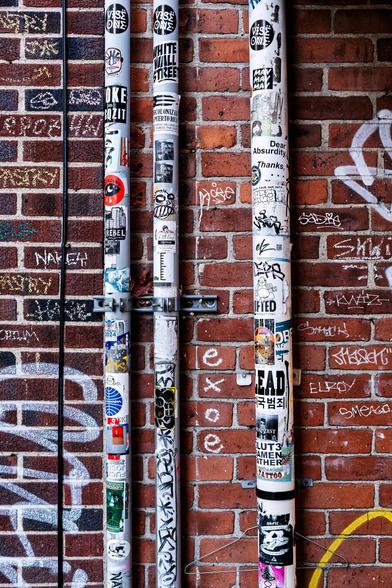 A color, daytime photograph, oriented vertically, showing three parallel, white PVC pipes mounted to a red brick wall in an alley. The pipes run top to bottom. The two narrower pipes are closer together and attached to a metal bracket. The wider pipe is set further to the right, and has a wire clothes hanger dangling from its bracket close to the bottom of the frame. The pipes are covered in stickers for bands, social/political groups or stances, some local establishments or seemingly random. The brick is covered with paint and chalk graffiti. The red bricks on the left edge - extending rightward until the first pipe, is a darker red than the rest of the wall.