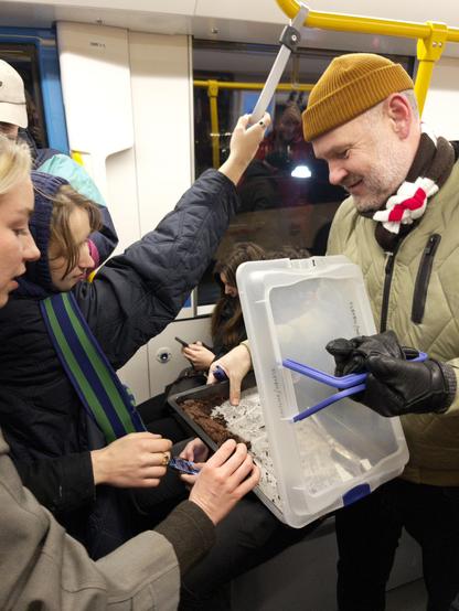 A photo of a man offering a tray of chocolate brownies to people on a busy tram.