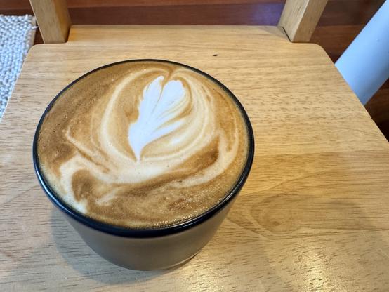 Small black coffee mug filled with a cappuccino sitting on a small wooden chair.